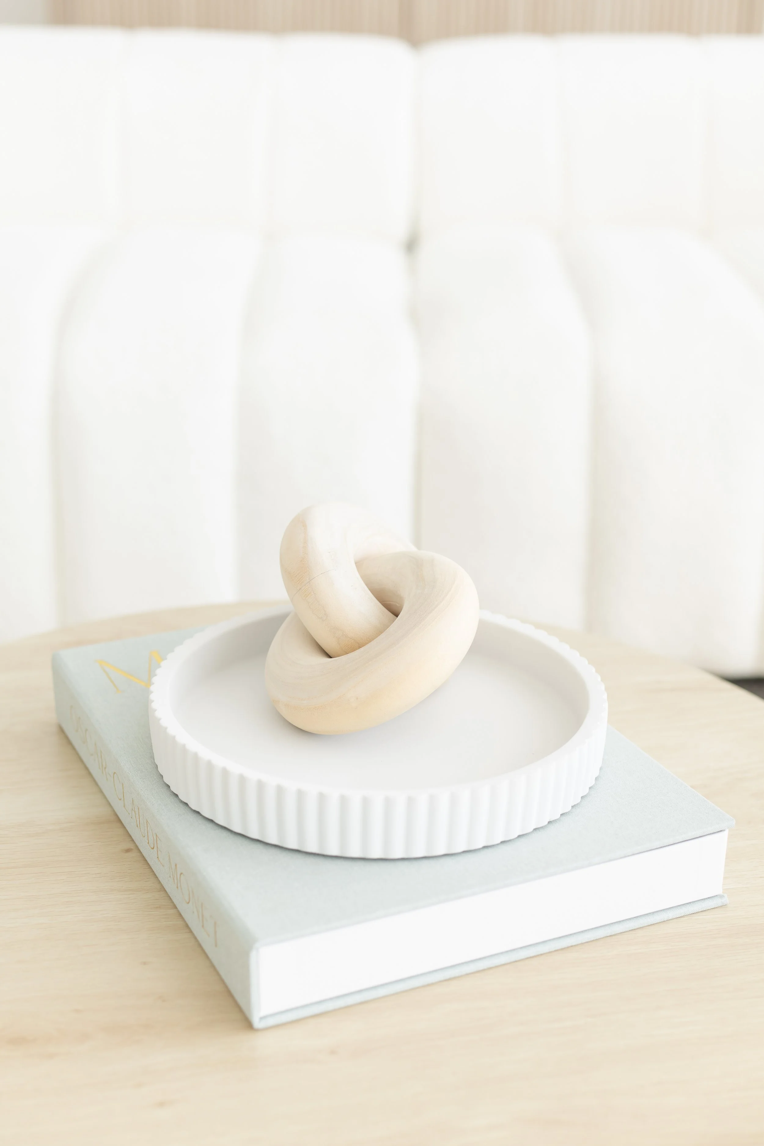 A white wooden knot sculpture placed on a round white tray on top of a white book with pastel blue edges and gold writing. The background is a white cushioned headboard.