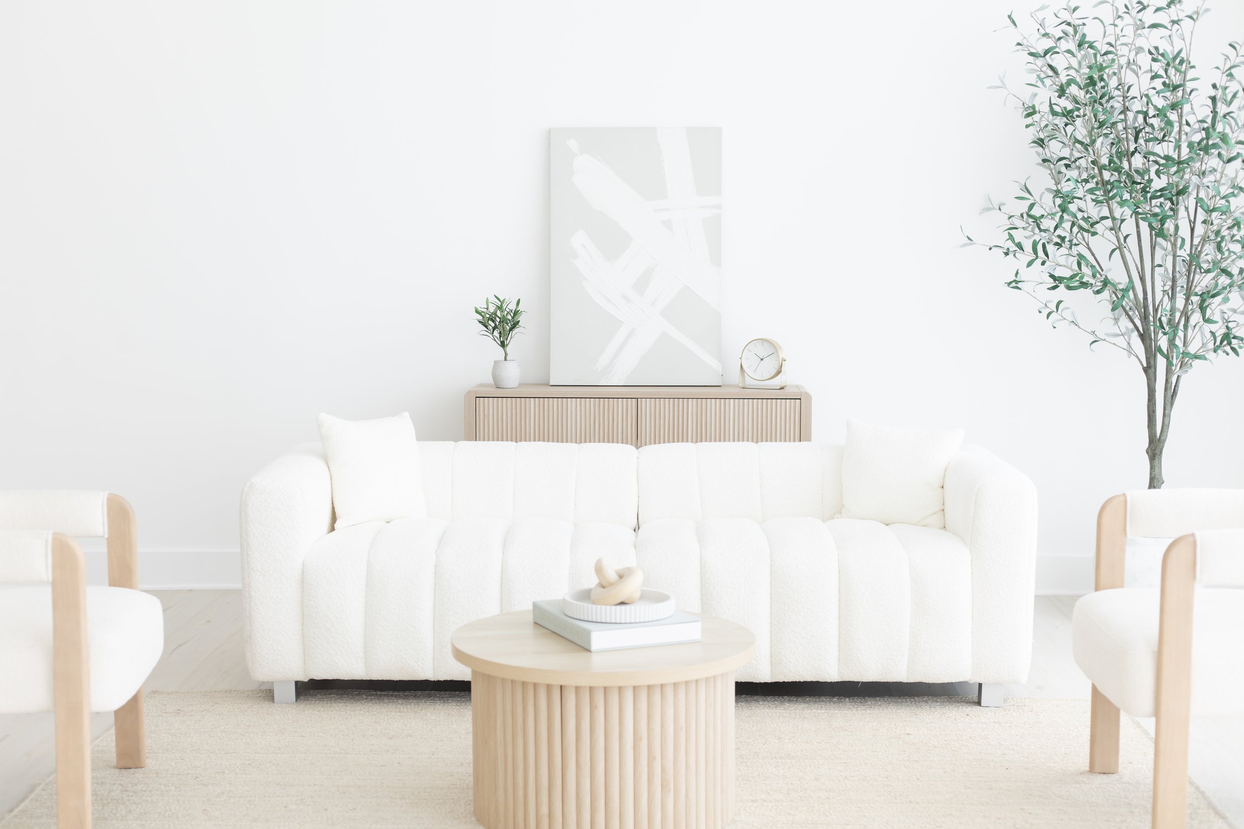 A minimalist living room with a white sofa, a round wooden coffee table, and a matching sideboard against a white wall. Decorative items include a small plant, abstract artwork, a clock, and a sculpture.