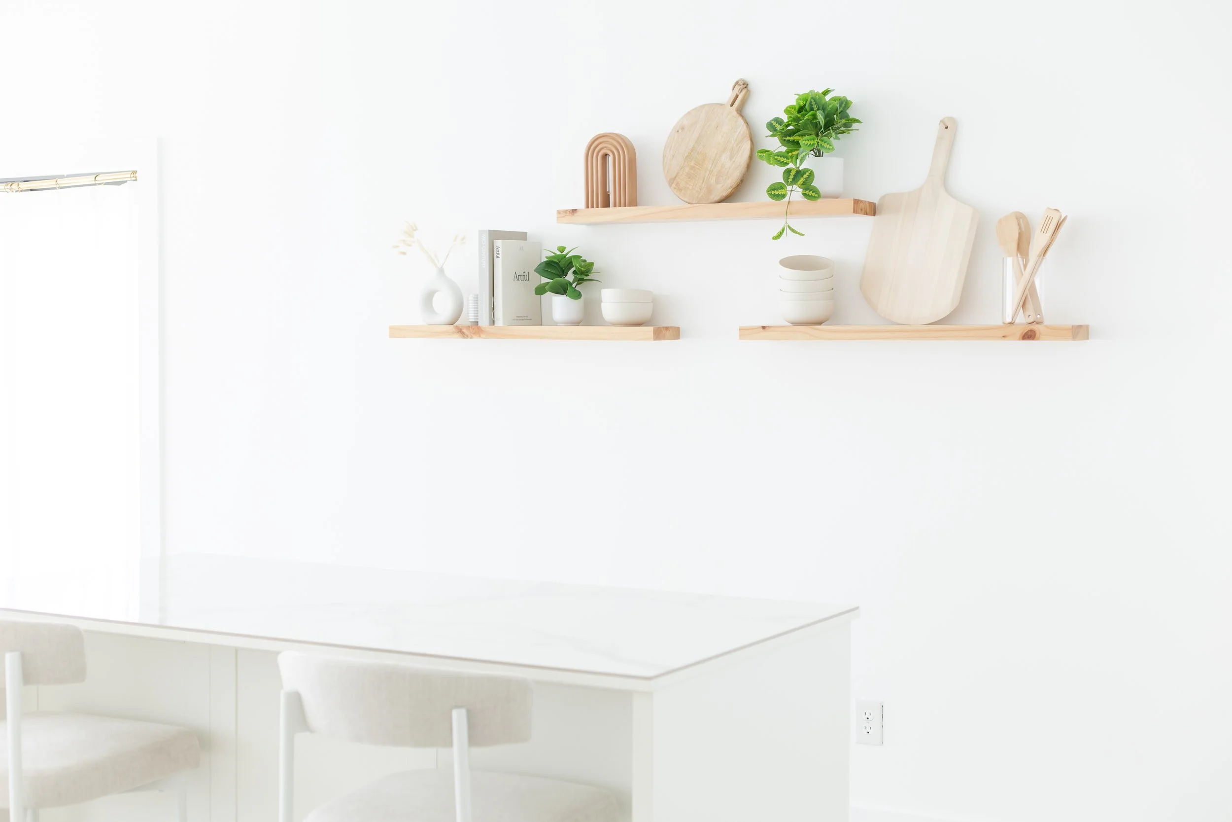 Minimalist kitchen with white wall, wooden shelves displaying green potted plants, cutting boards, and bowls, with a glimpse of a white table and chair.