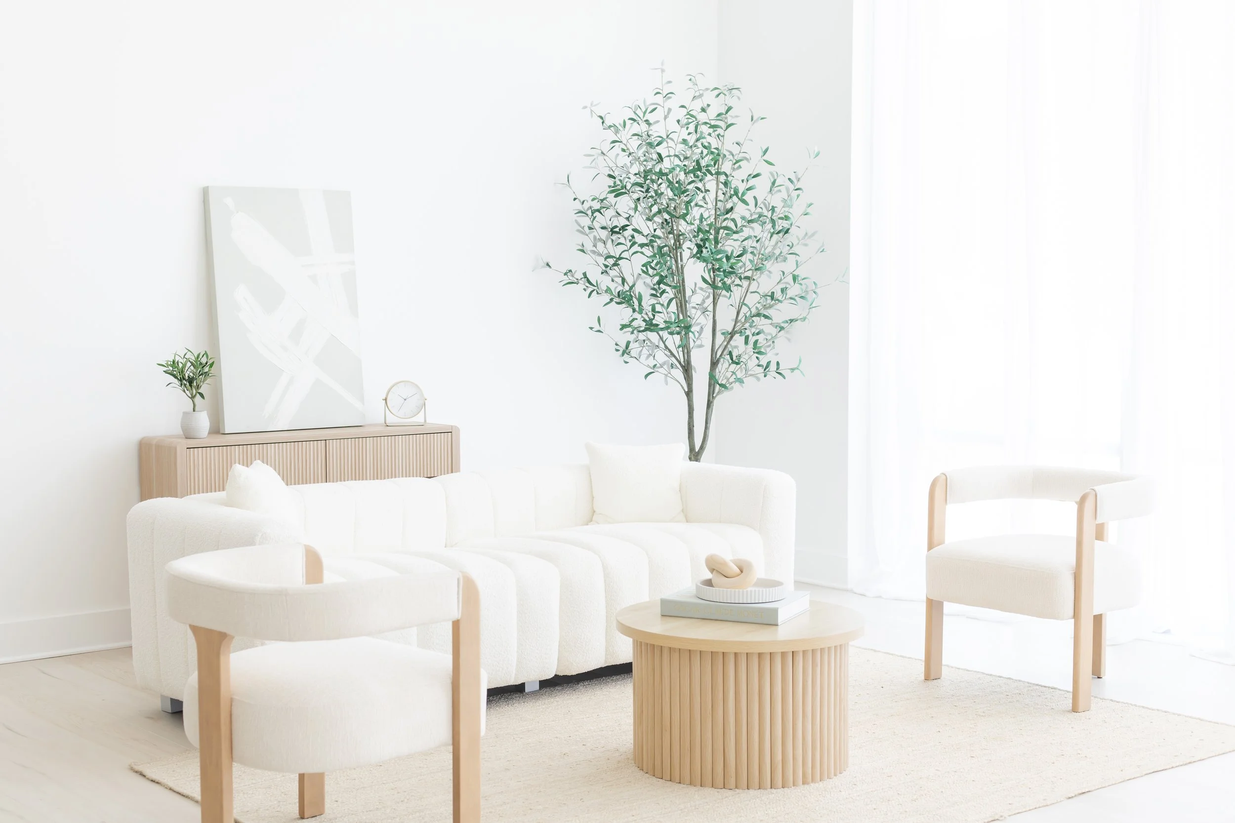 Bright living room with white furniture, a wooden side table, a sofa with pillows, a large potted tree, and a wall decorated with a clock, a small potted plant, and a large abstract painting.