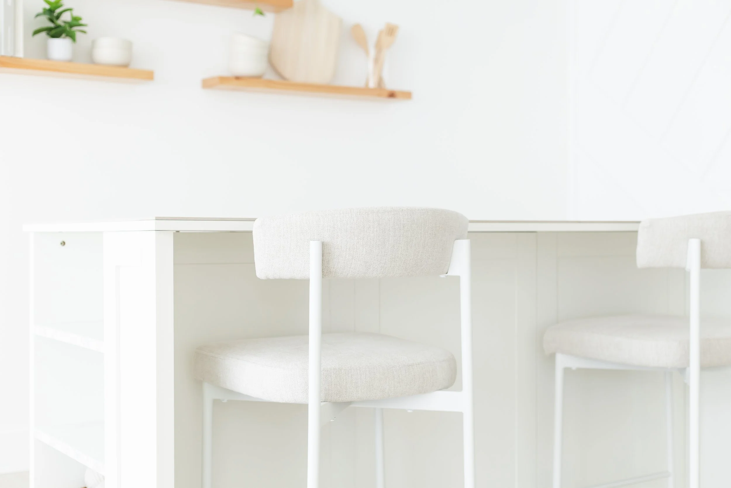 A white desk with a matching white chair in a bright, minimalist room. Two wooden floating shelves on the wall hold potted plants and wooden kitchen utensils, with a white wall and window in the background.