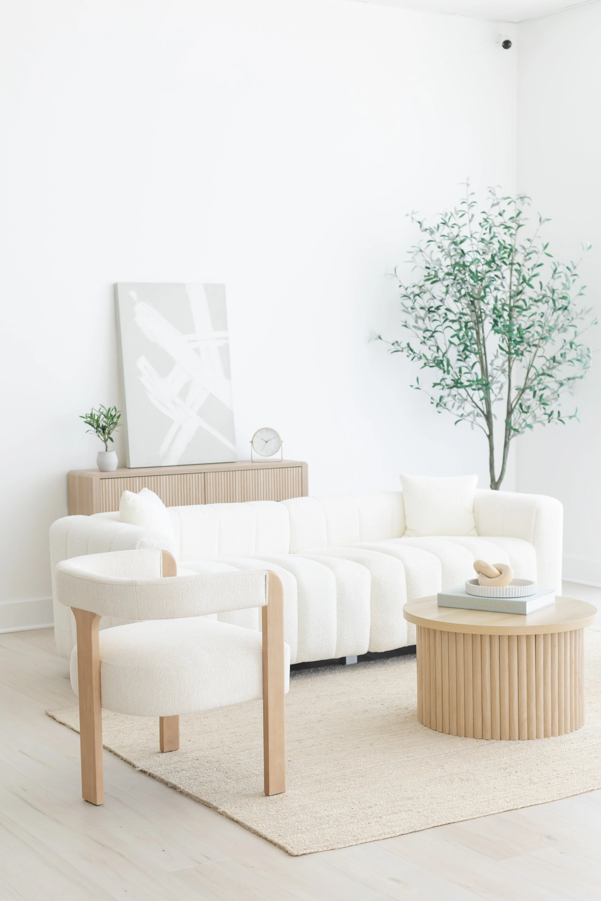 A minimalist living room with a white sofa, a light wood armchair, round light wood coffee table, large potted plant, abstract wall art, small clock, books, and neutral-colored decor on a beige rug.