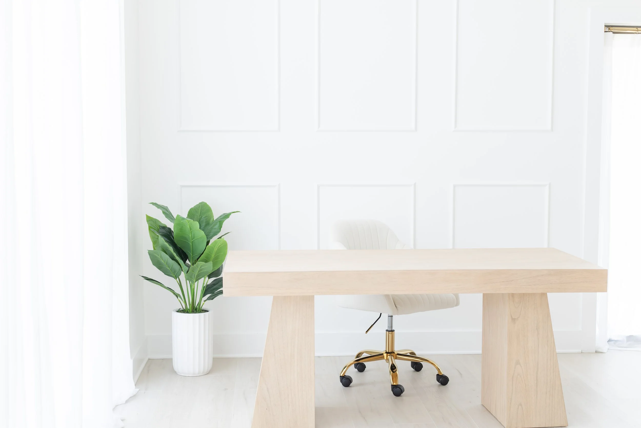 A modern minimalist office space with light wood desk, beige office chair on wheels, green potted plant in white pot, white wall with decorative panels, and sheer white curtains.