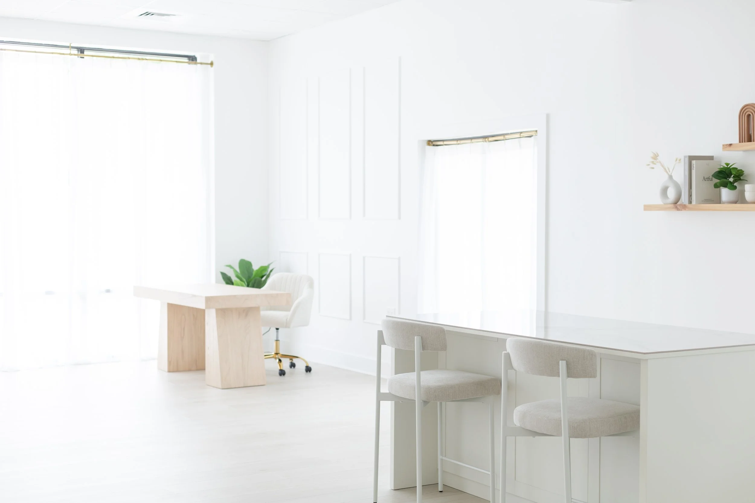 Bright, minimalist white interior with a dining or workspace area, featuring a white table, white chairs, a floating shelf with a plant and decorative items, and large windows with sheer curtains letting in natural light.