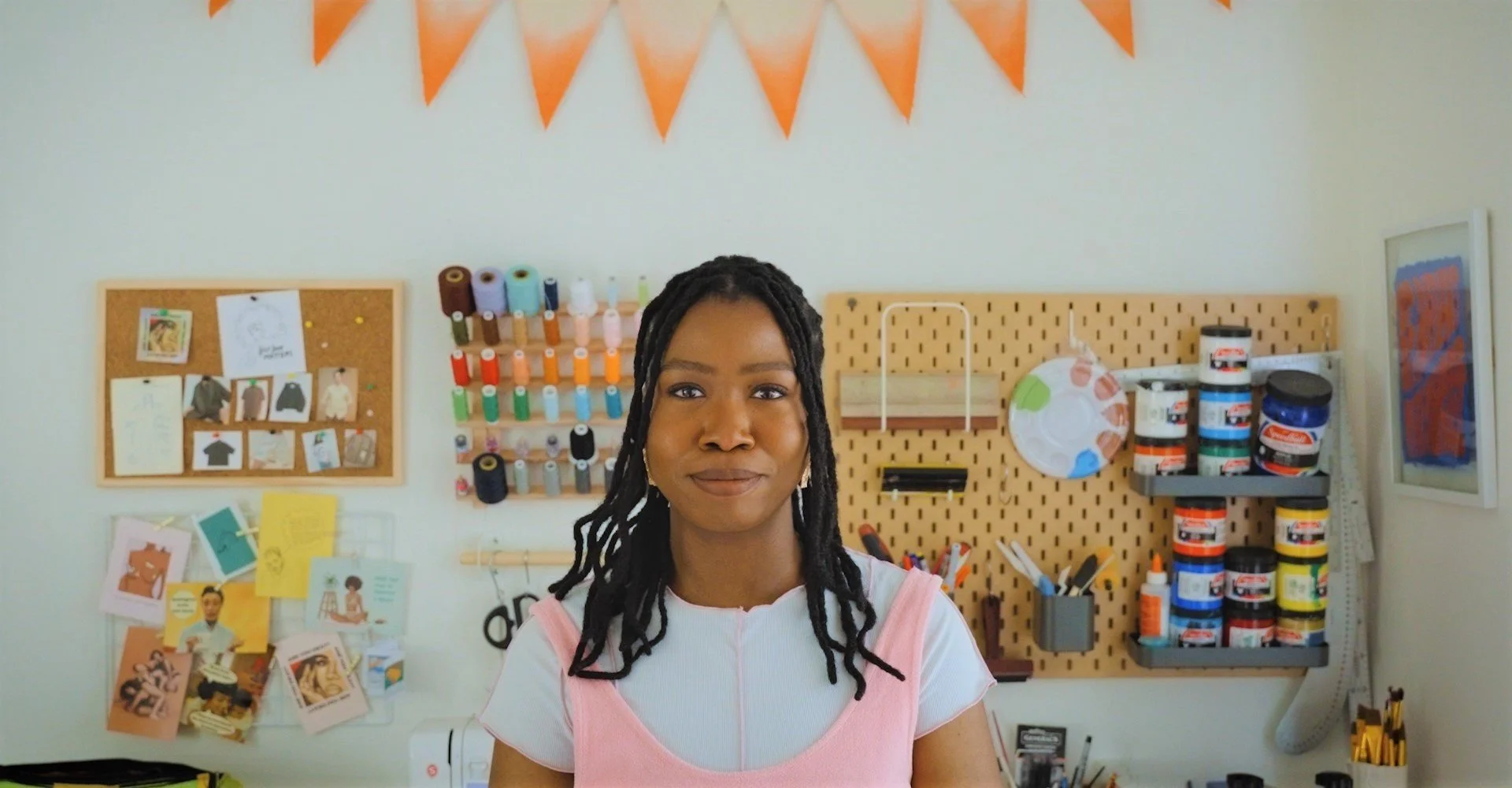 Une femme souriante pose devant un mur rempli d'articles d'artisanat et de fournitures créatives, dont des bobines de fil, des pots de peinture et des photos et dessins accrochés au mur.
