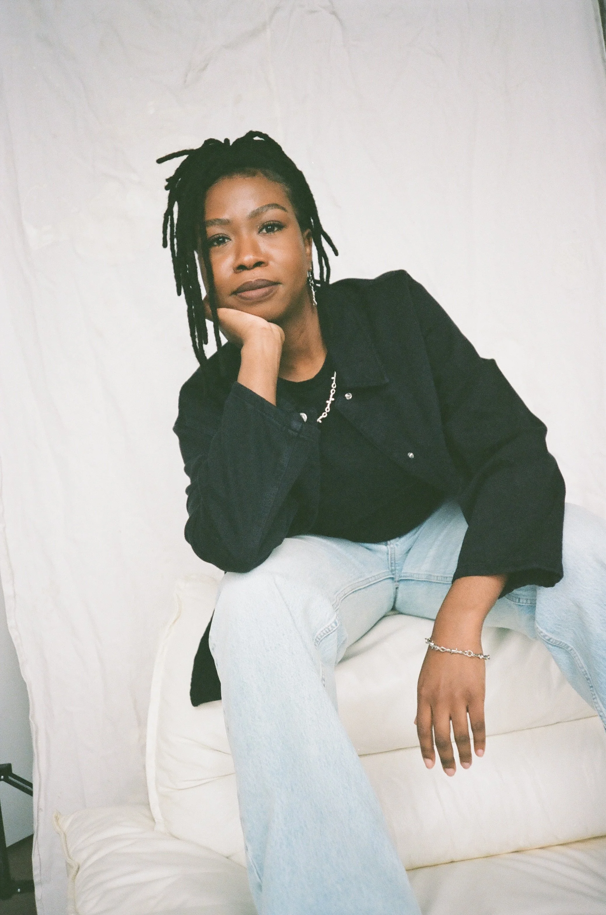 Une femme avec des dreadlocks assise sur un canapé blanc, portant une veste noire et un jean clair, posant avec la tête appuyée sur la main contre un mur blanc.