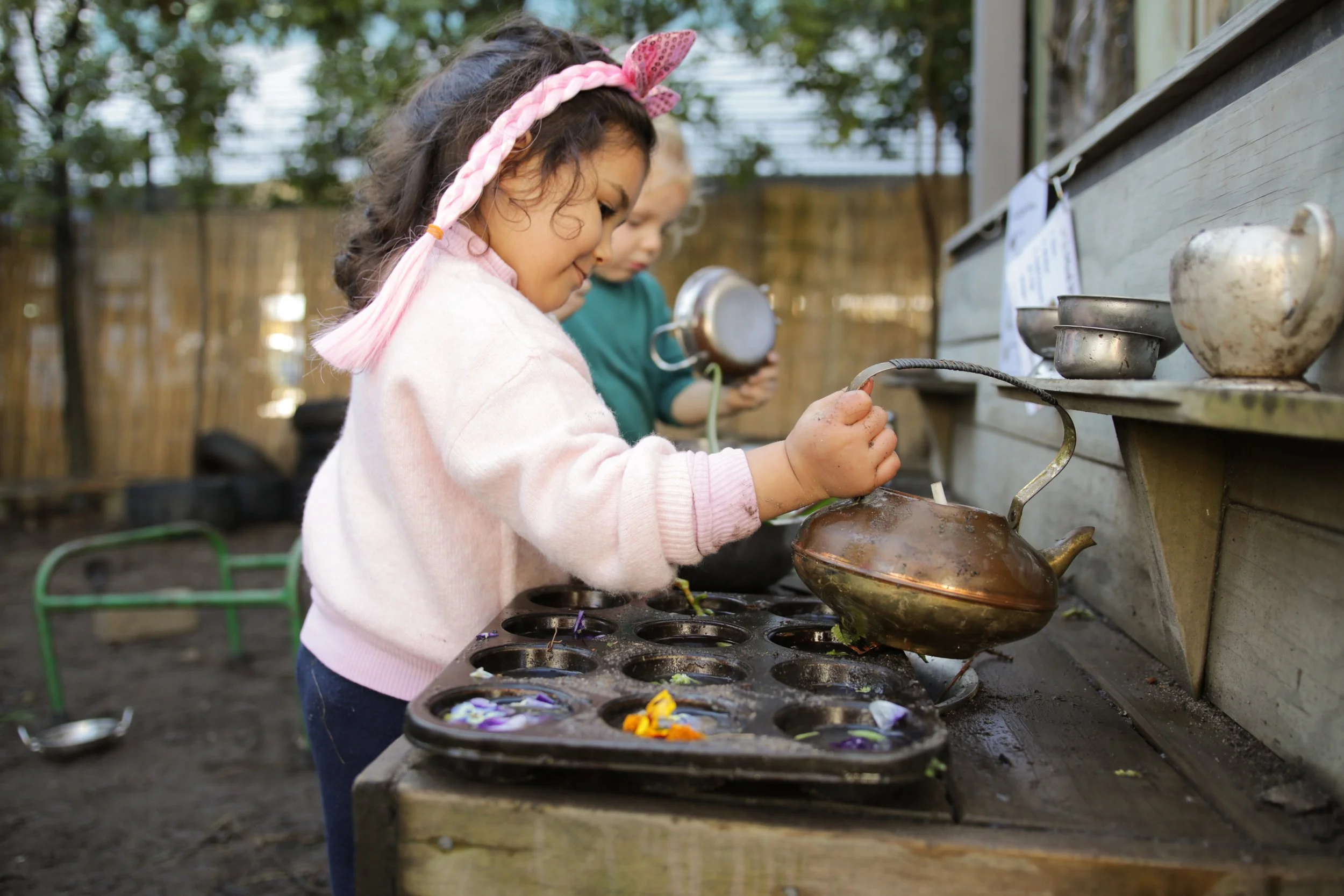 Two young girls, one with curly hair and a pink headband, are planting flowers in a wooden outdoor grow station with metal bowls filled with colorful flower petals and small plants.