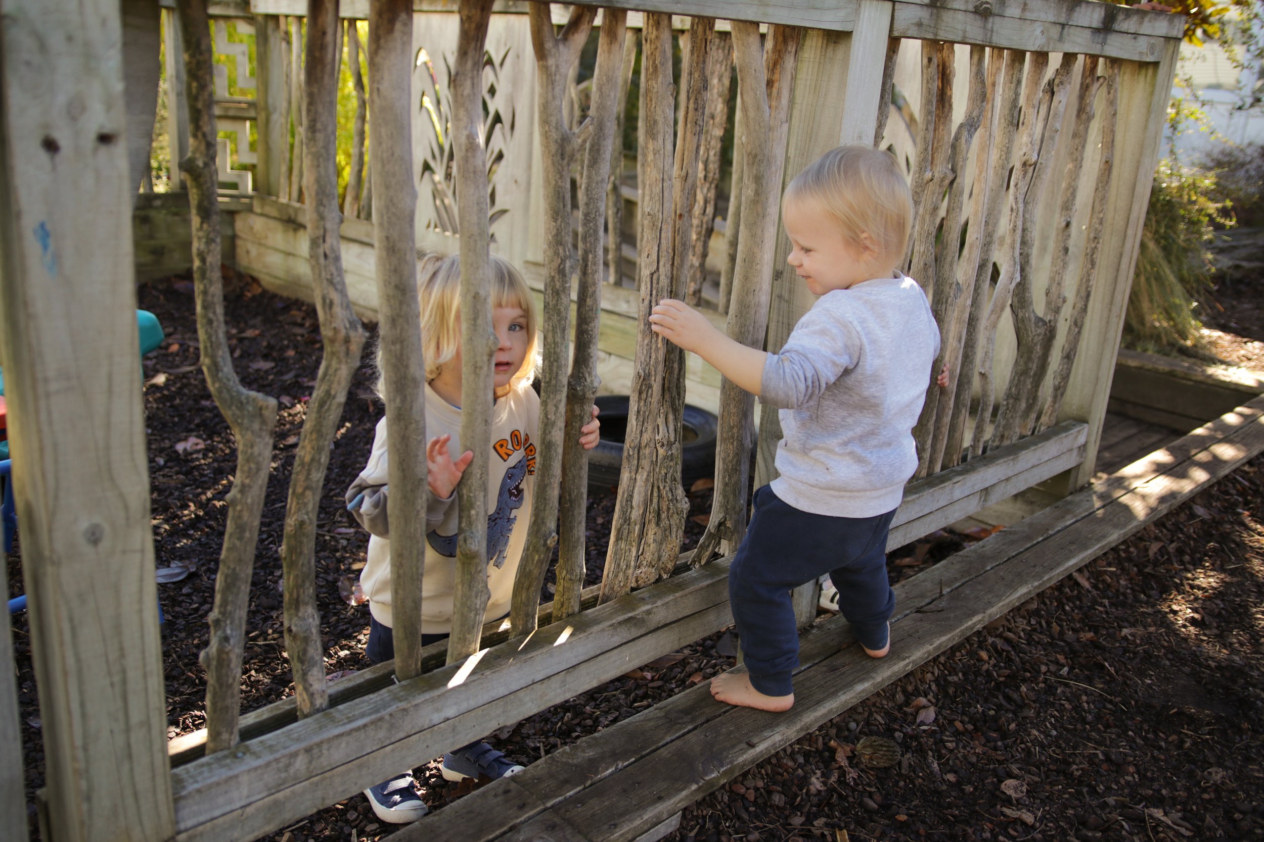 Two young children, a boy and a girl, playing behind a wooden garden fence with vertical slats. The boy with blonde hair is standing barefoot, wearing a light gray long-sleeve shirt and dark pants, smiling while touching the fence. The girl, with blonde hair and a light-colored shirt with a dinosaur graphic, is peering through the slats. Sunlight illuminates the scene, and there are trees and garden soil visible.