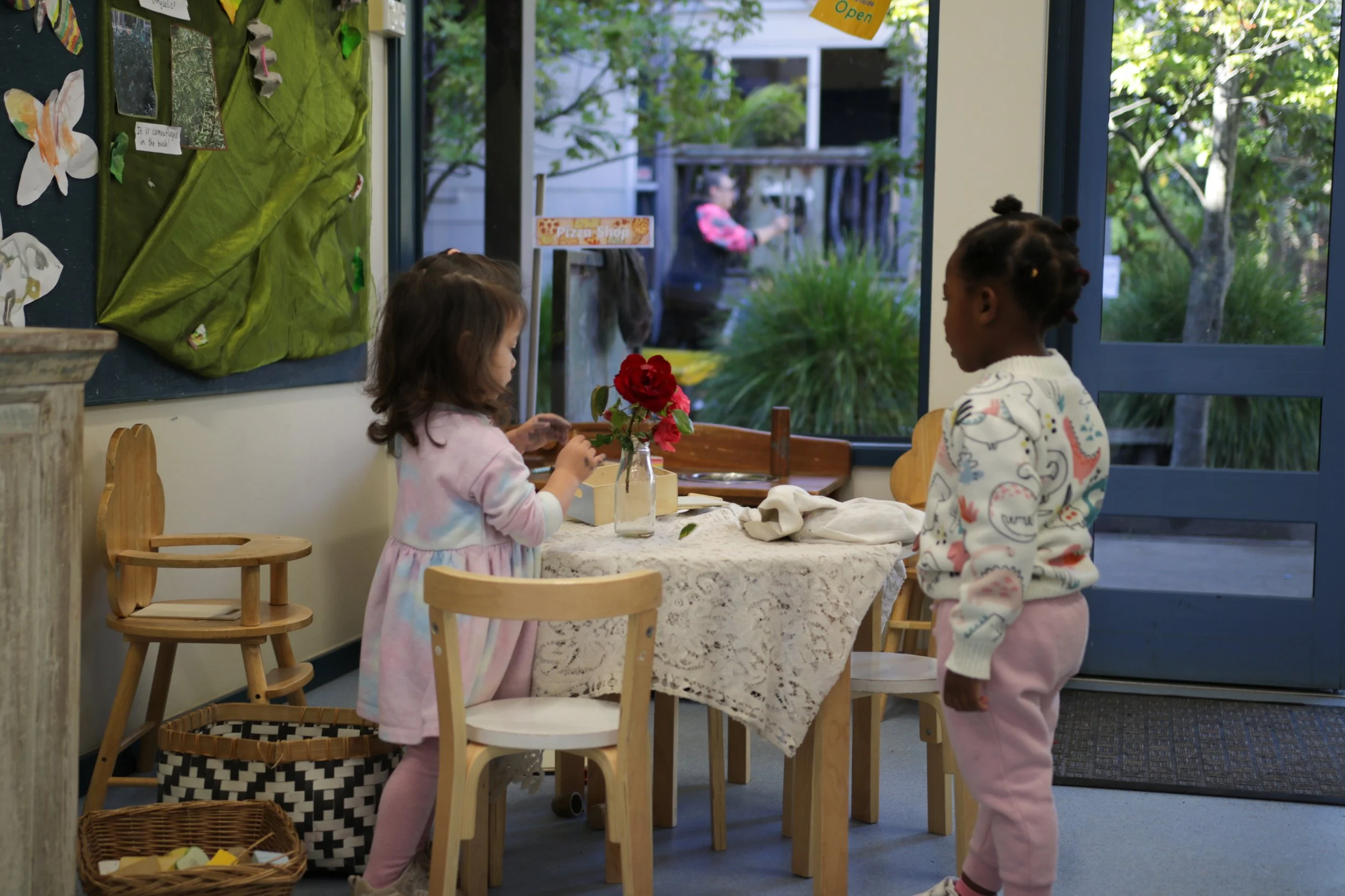 Two young girls with pigtails and a girl with brown hair in a pink dress, standing and sitting at a table inside a room, with a glass jar holding red flowers on the table. There is a bulletin board with children’s artwork and a window showing an outdoor garden with trees and a person passing by.