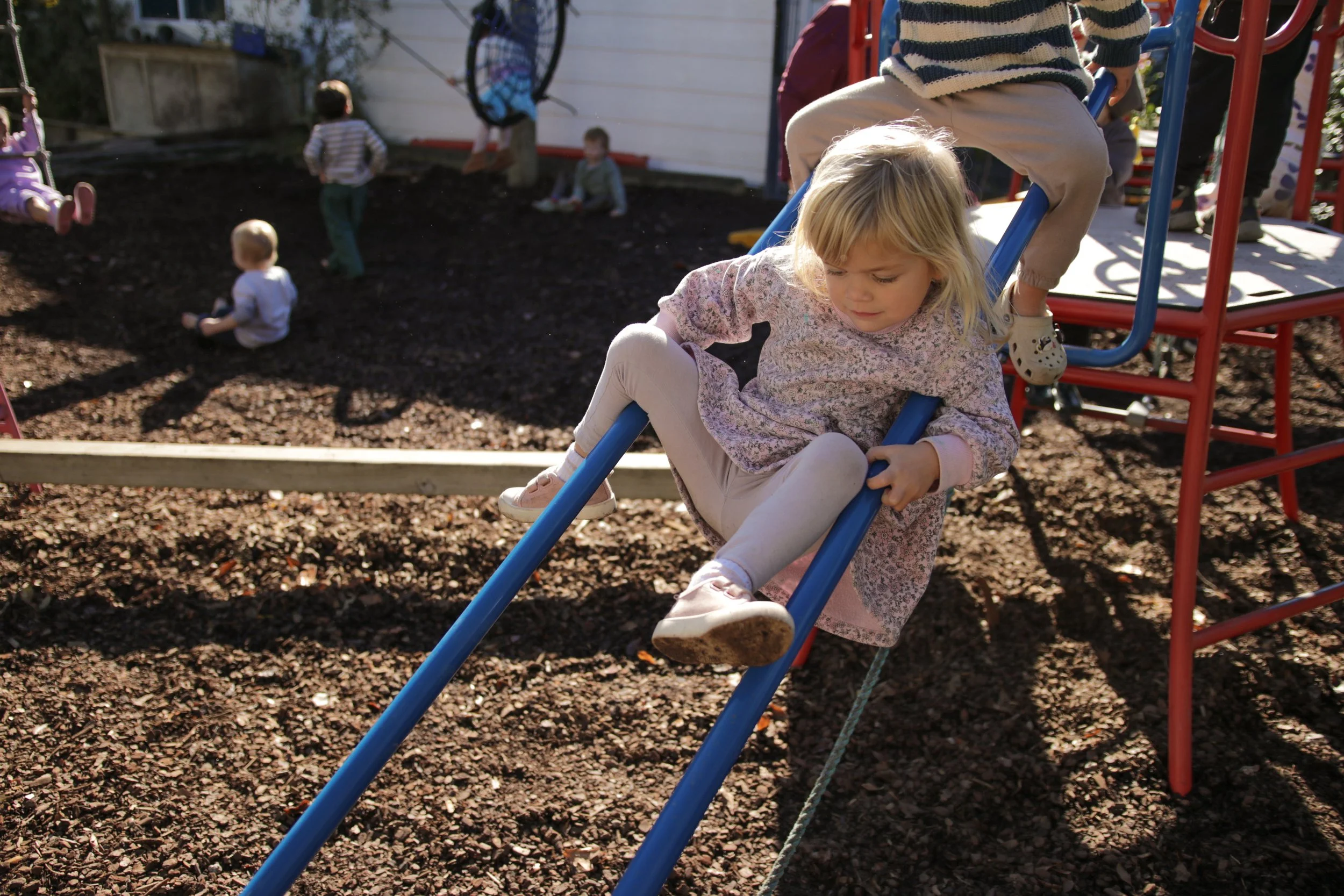 Young girl with blonde hair playing on a playground slide, other children in the background, outdoors on a sunny day.