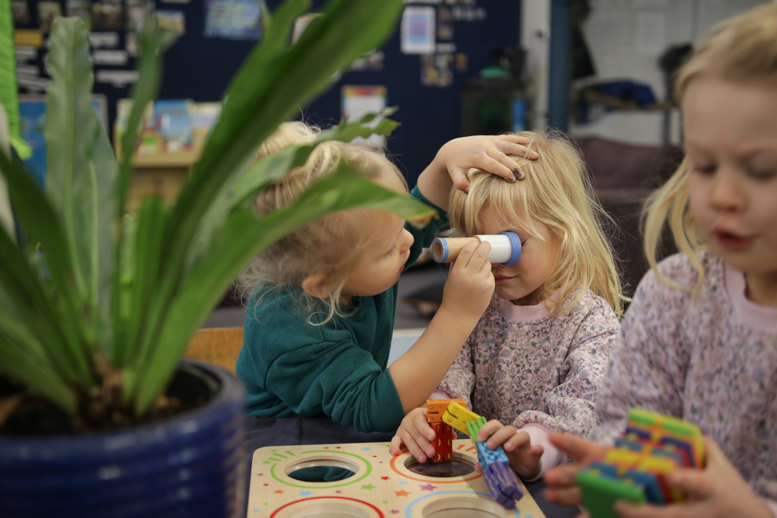 Children playing a game with colorful blocks at a table in a classroom. A potted plant is in the foreground, partially blocking the view.