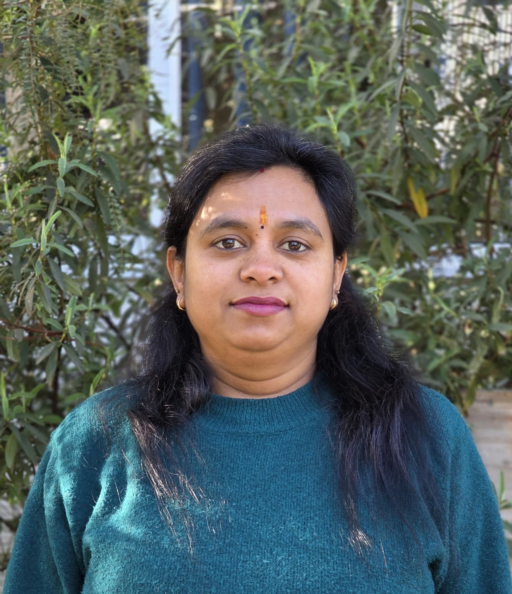 A woman with dark hair and a red and yellow bindi, wearing earrings and a teal sweater, standing in front of green foliage.