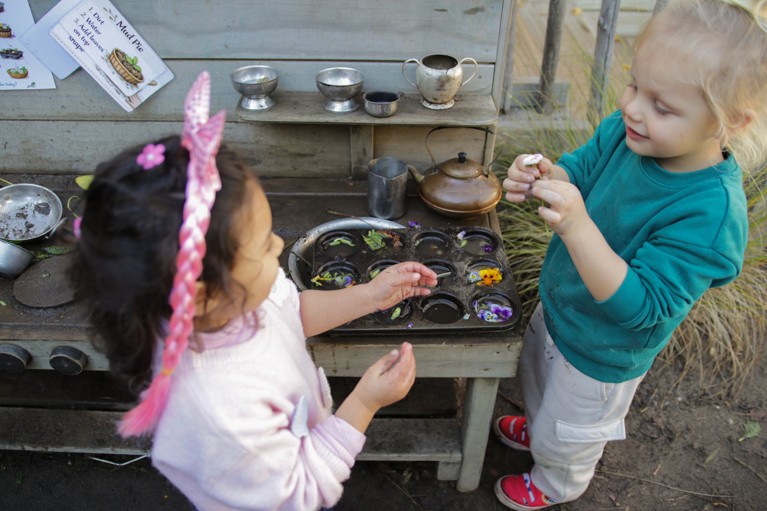 Two young children engaging in a garden activity with mud and flowers, near a wooden outdoor kitchen setup with metal cups, a teapot, and instructions for making mud pie.