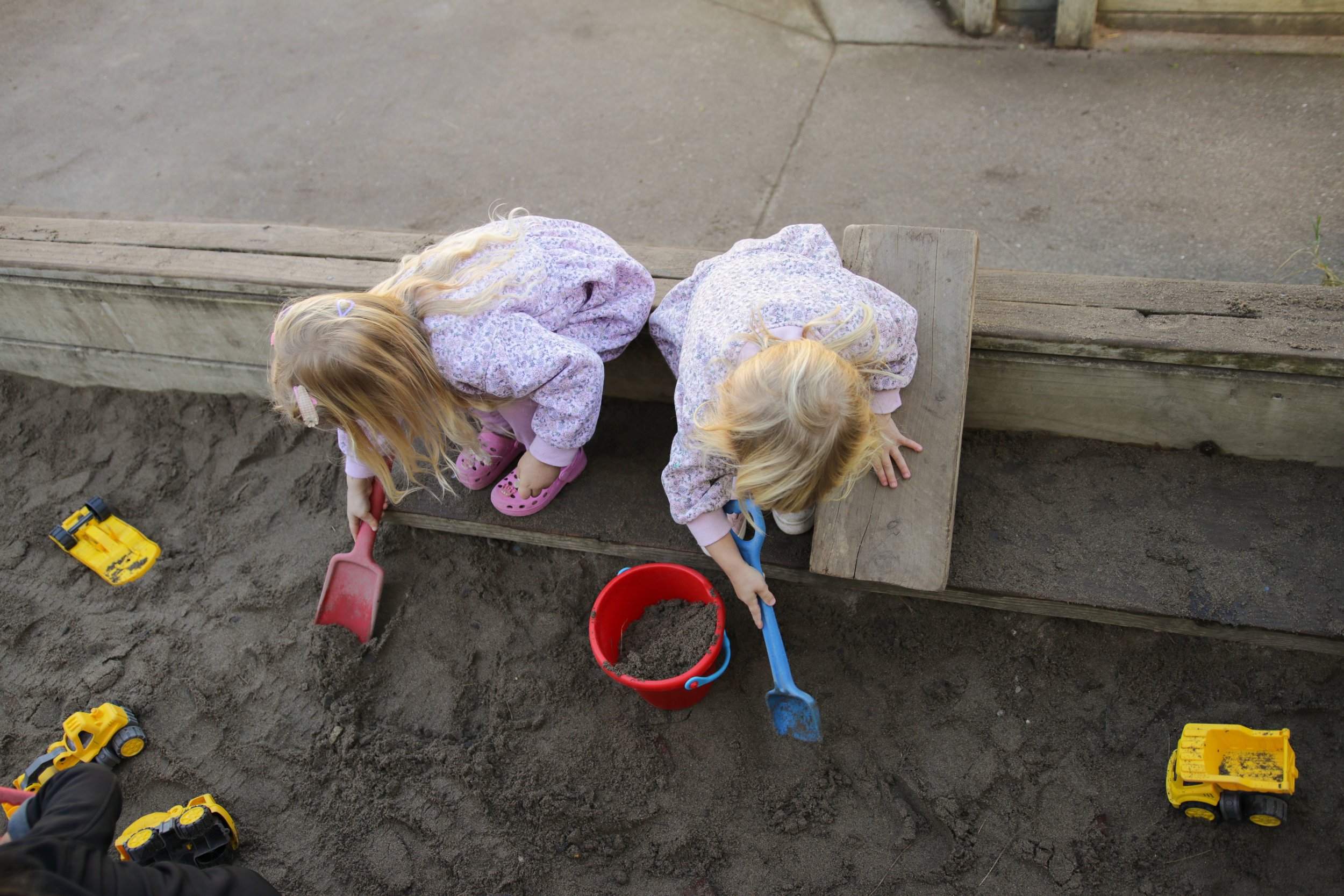 Two young girls playing in a sandbox, scooping sand into a red bucket with shovels, surrounded by toy construction vehicles, in an outdoor play area.