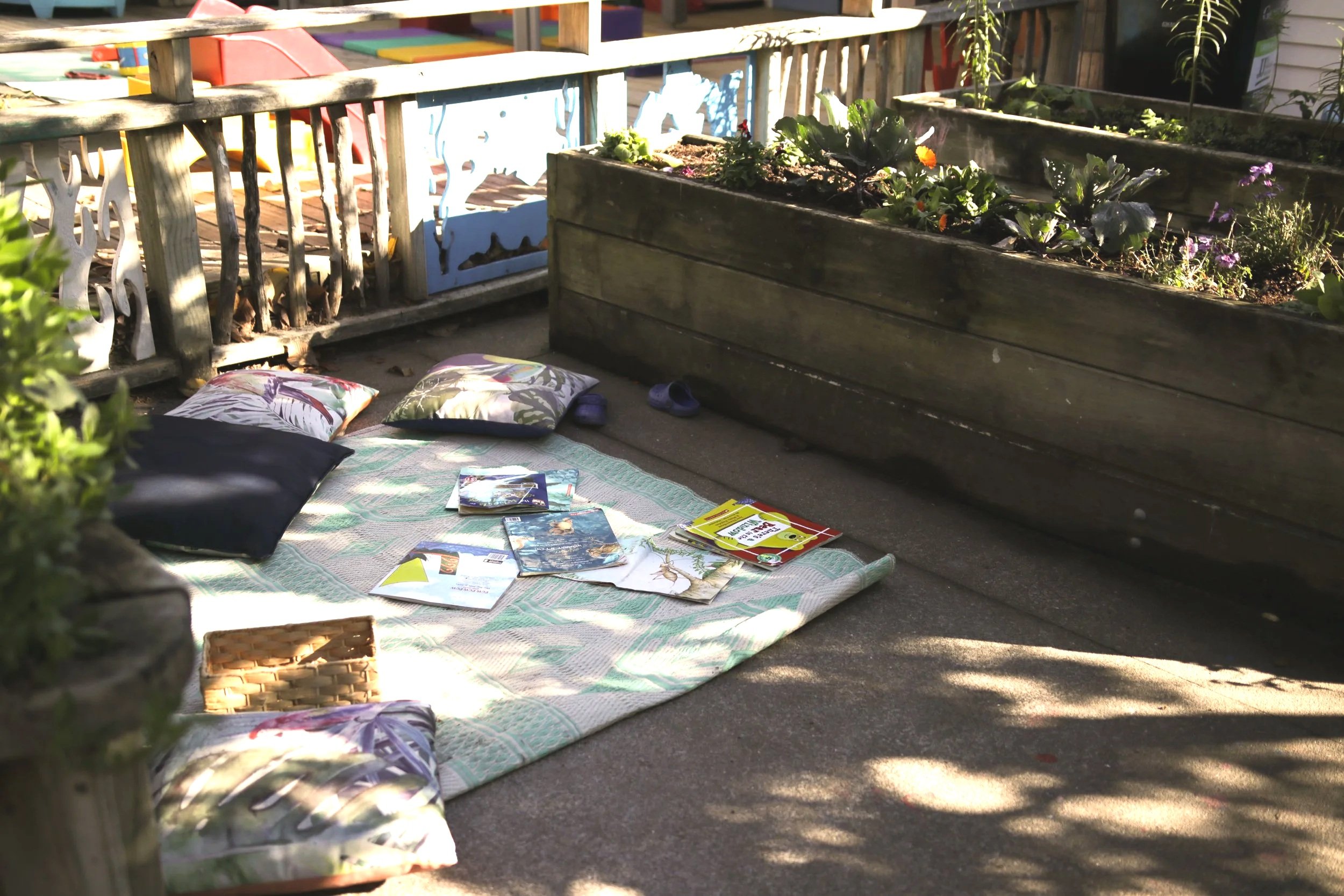 A mantle shelf with flowers and a garden raised bed with plants, and a mat with pillows, magazines, and newspapers on the ground outside in sunlight.