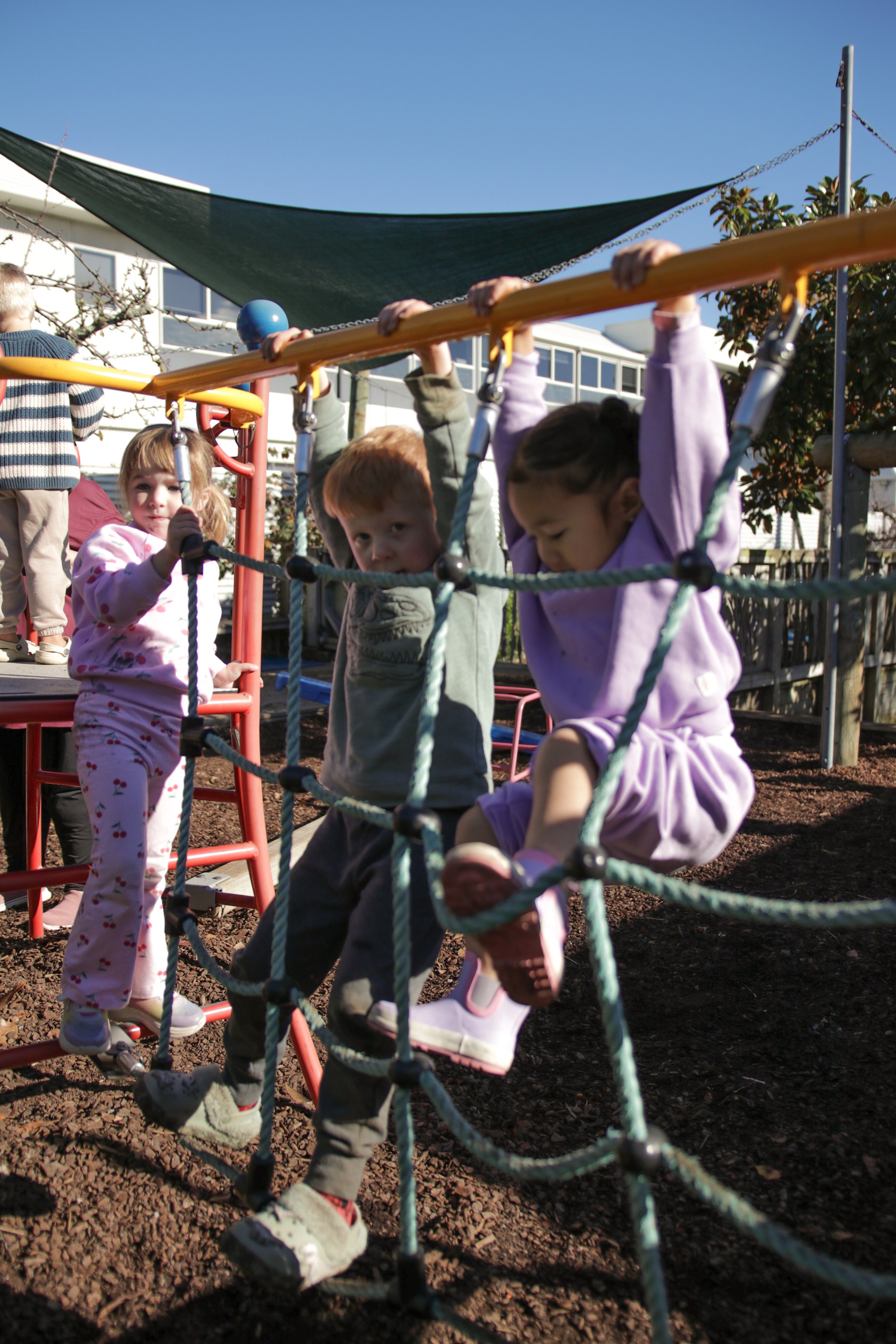 Children playing on a rope climbing structure in a playground with a building and trees in the background under a clear blue sky.