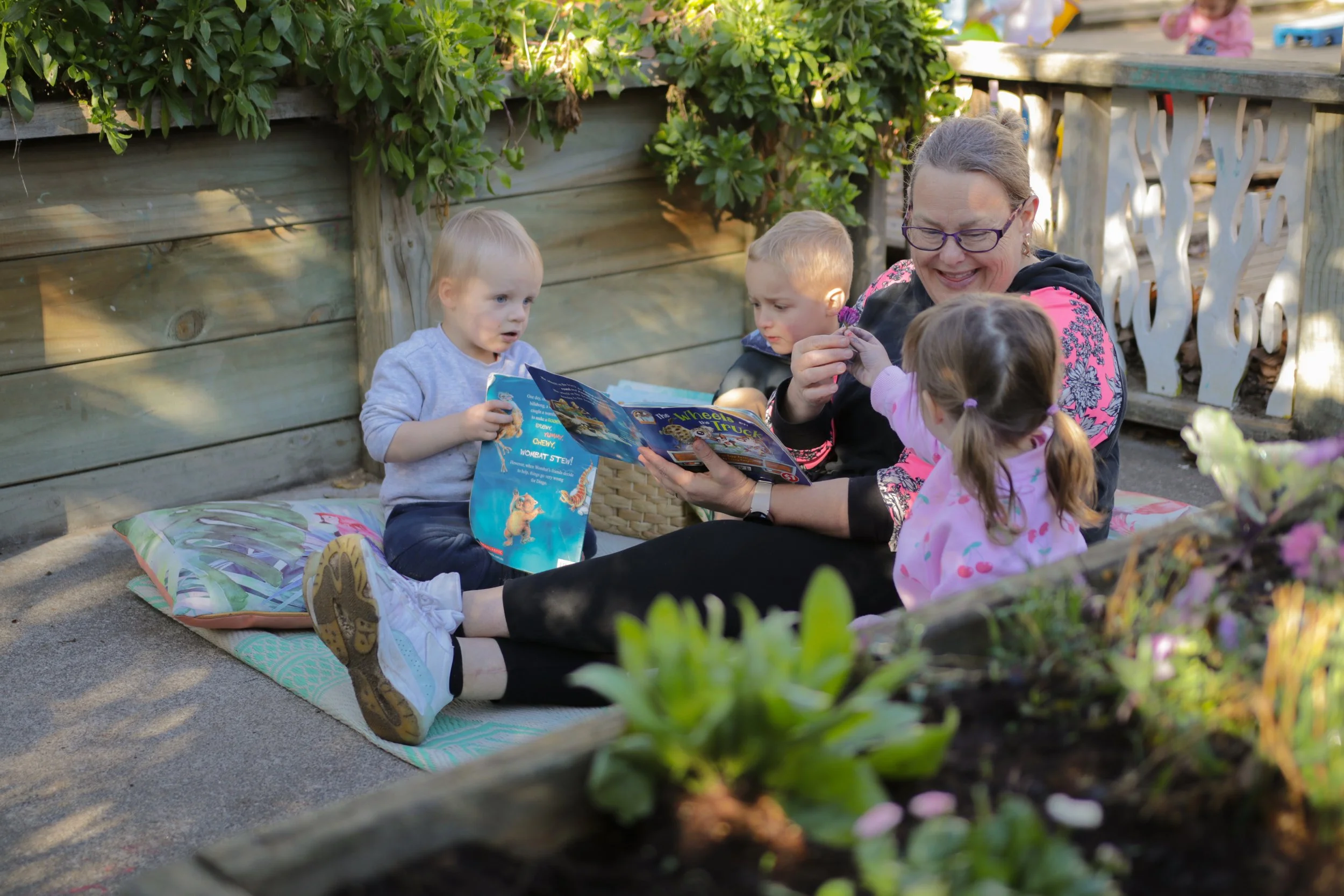 A woman and three children sitting on a blanket outdoors, reading a colorful children's book with a garden fence in the background.