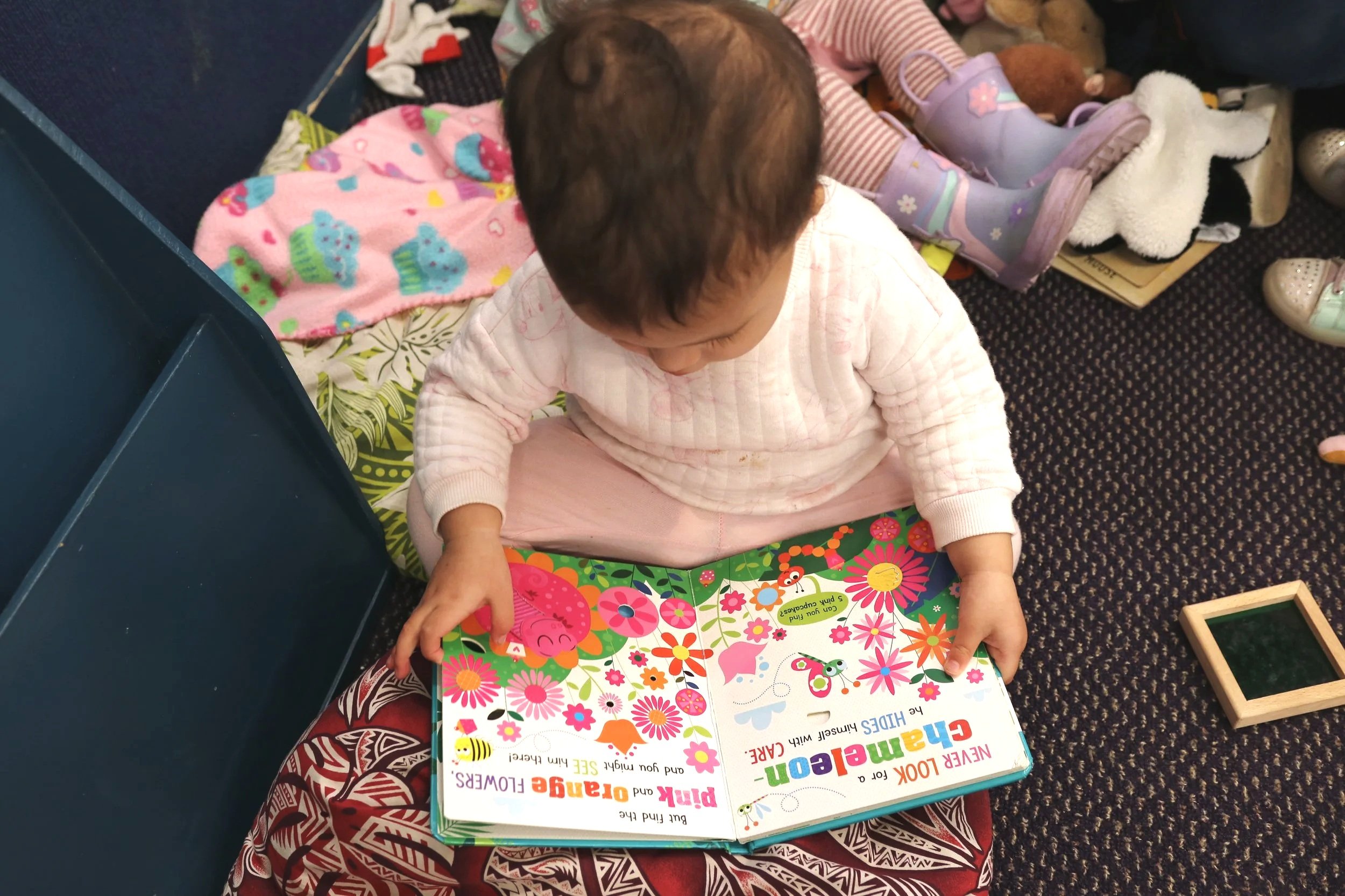 A young child sitting on a colorful blanket, looking at a large, vibrant children's book with bright flowers and animals on the cover, surrounded by toys and shoes.