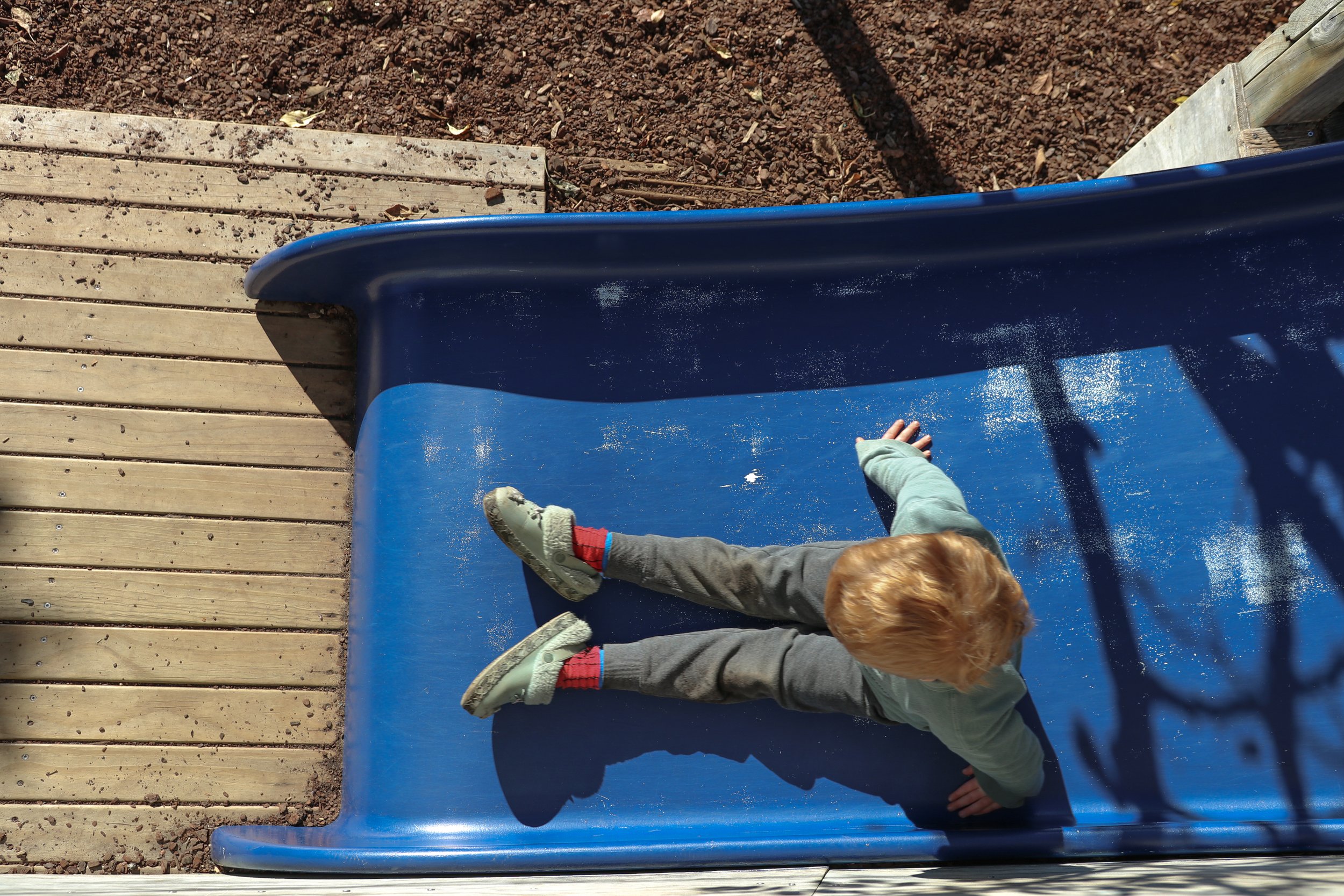 Child climbing down a blue slide at a playground, with wooden planks on the left and dirt on the ground.