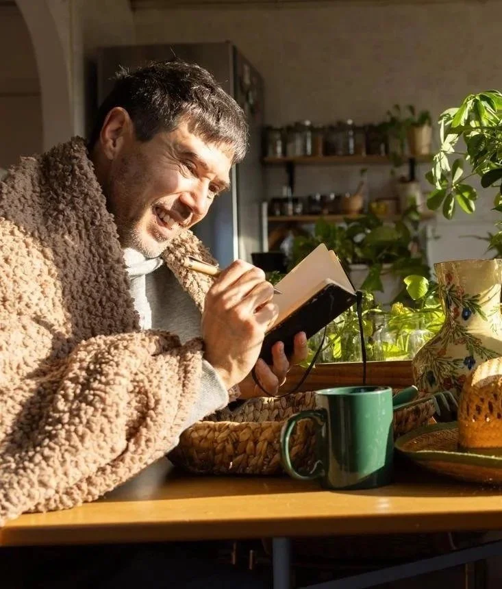 A man sitting at a table, smiling and writing in a notebook, with a warm fleece coat, mug, and decorative vase on the table, in a cozy kitchen with sunlight.
