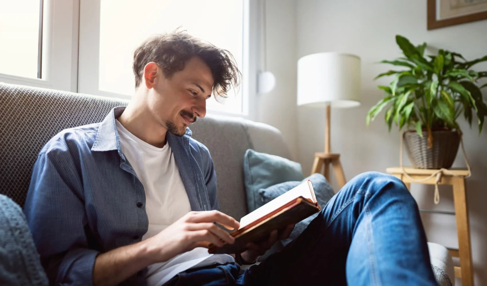 A young man with brown hair, plaid shirt, and white T-shirt sitting on a couch, smiling while reading a book in a bright living room with a large window, houseplants, and a lamp.
