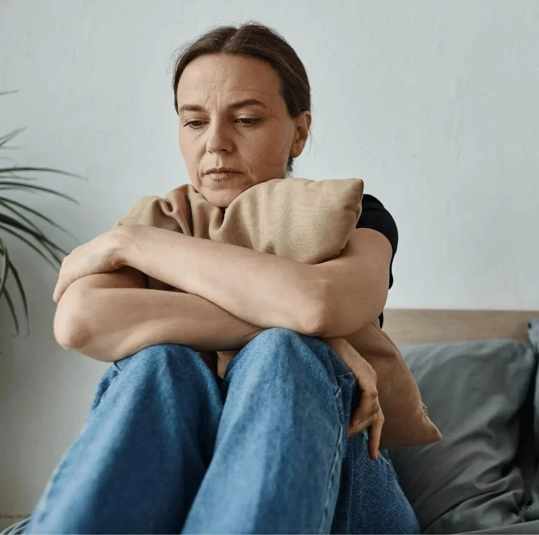A woman sitting on a bed, hugging a pillow and looking down with a sad expression.