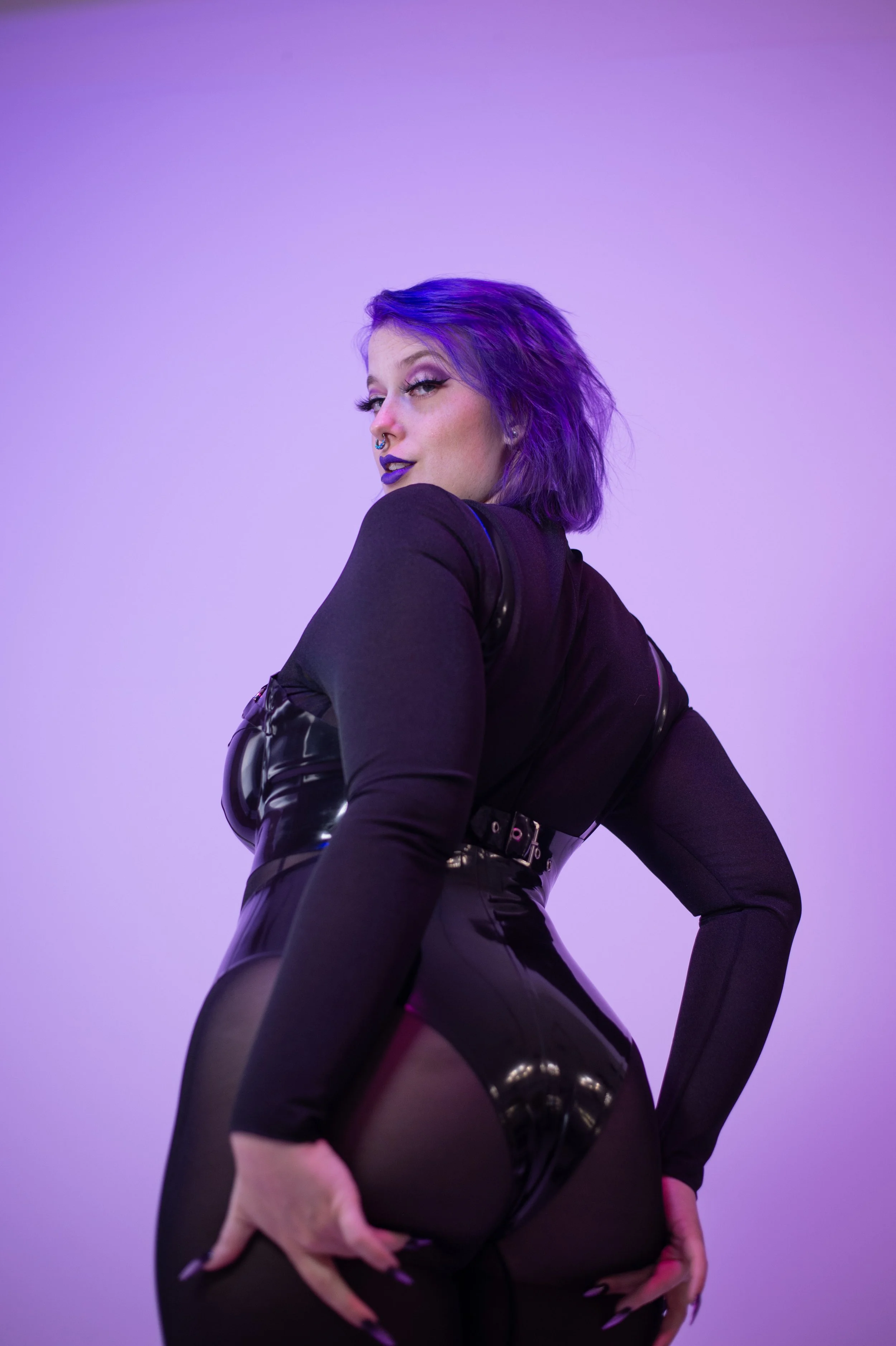 Goddess Nyx cups her round butt, looking back at the camera. A purple background meshes well with her purple hair as she looks right at you