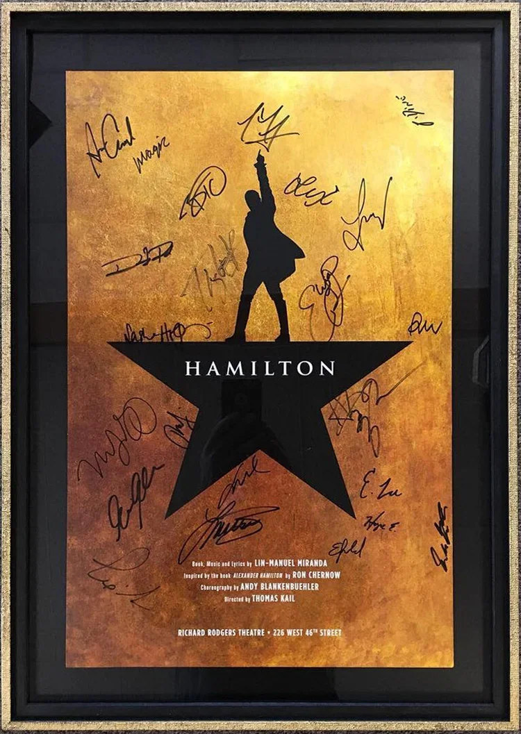 Signed poster of the musical Hamilton, featuring a silhouette of Alexander Hamilton holding a flag, against a gold background, enclosed in a black frame.  custom framing brookline pictureplacebrookline.com