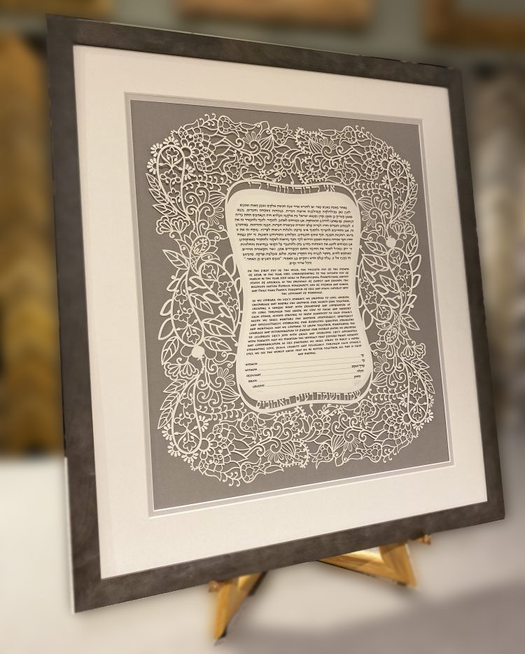 Framed decorative document with intricate floral cut-out border, containing text in Hebrew, displayed on a small easel.