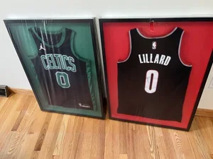 Two framed basketball jerseys, one of the Boston Celtics with number 0, and the other of Lillard, also with number 0, displayed on a wooden floor.  custom framing brookline pictureplacebrookline.com