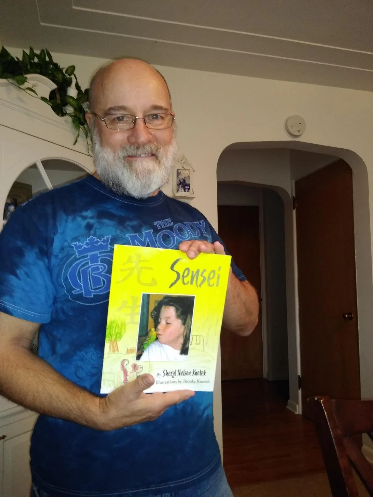 A man with glasses, a gray beard, and a blue T-shirt is holding a yellow book titled 'Sensei' by Sheryl Nelson Knotek with illustrations by Brooke Knotek, and a cover image of a woman with a yellow bird. He is standing in a room with a white wall, a partially open wooden door, a smoke detector, and a decorative plant.