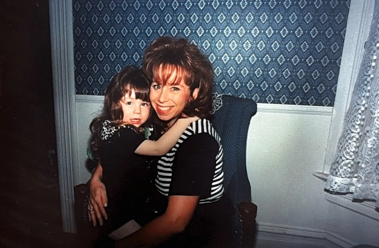 A woman with shoulder-length reddish hair and a young girl with dark brown hair and bangs are sitting together in a room with patterned blue wallpaper and lace curtains. The woman is smiling widely while the girl appears slightly concerned or confused. They are hugging each other, and the woman is wearing a black shirt with a black and white striped vest.