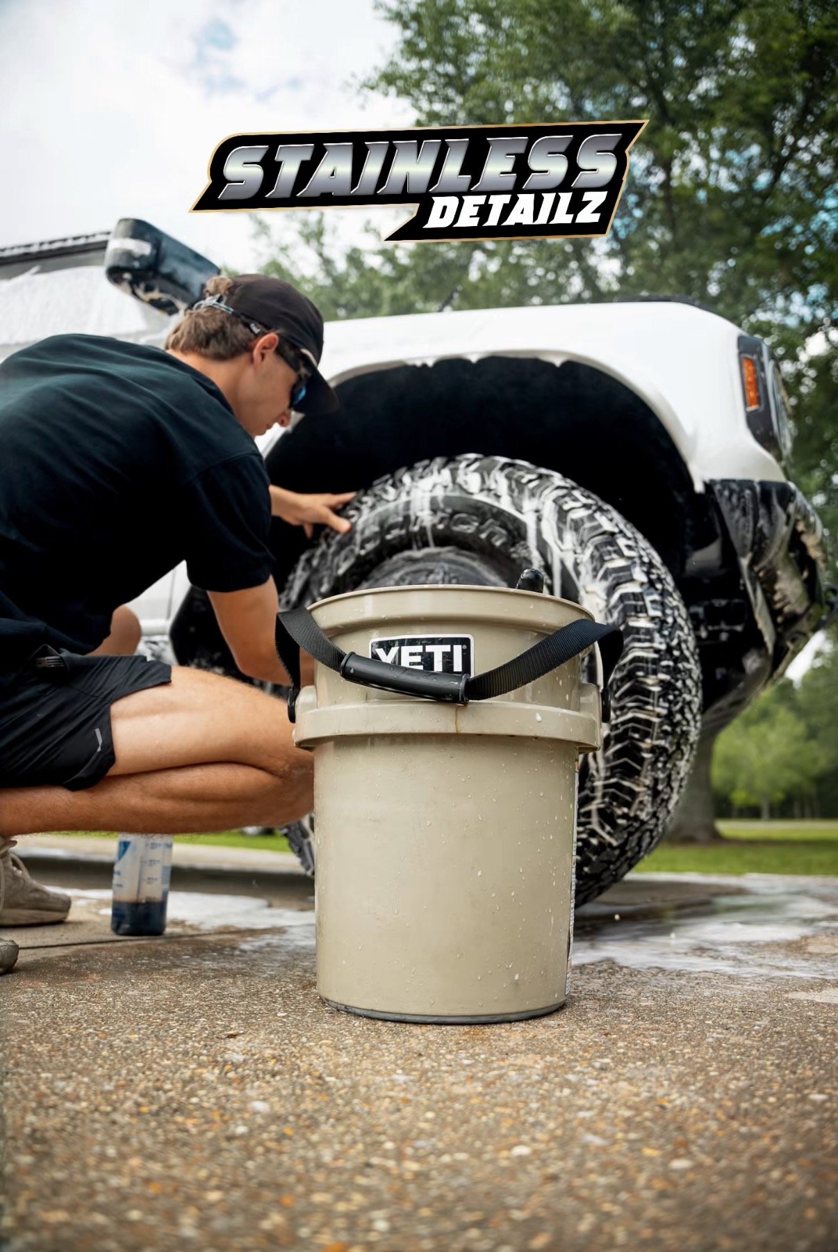 A person washing a white vehicle with a spray gun, with a bucket of water labeled 'YETI' nearby, outdoors with trees in the background.