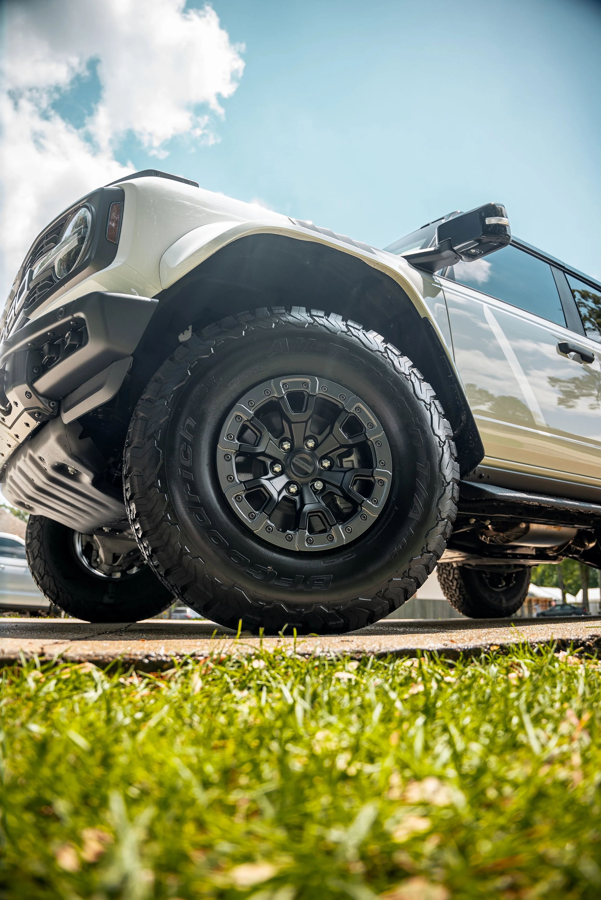 Close-up of a white off-road vehicle with large tires parked on grassy ground under a blue sky with clouds.