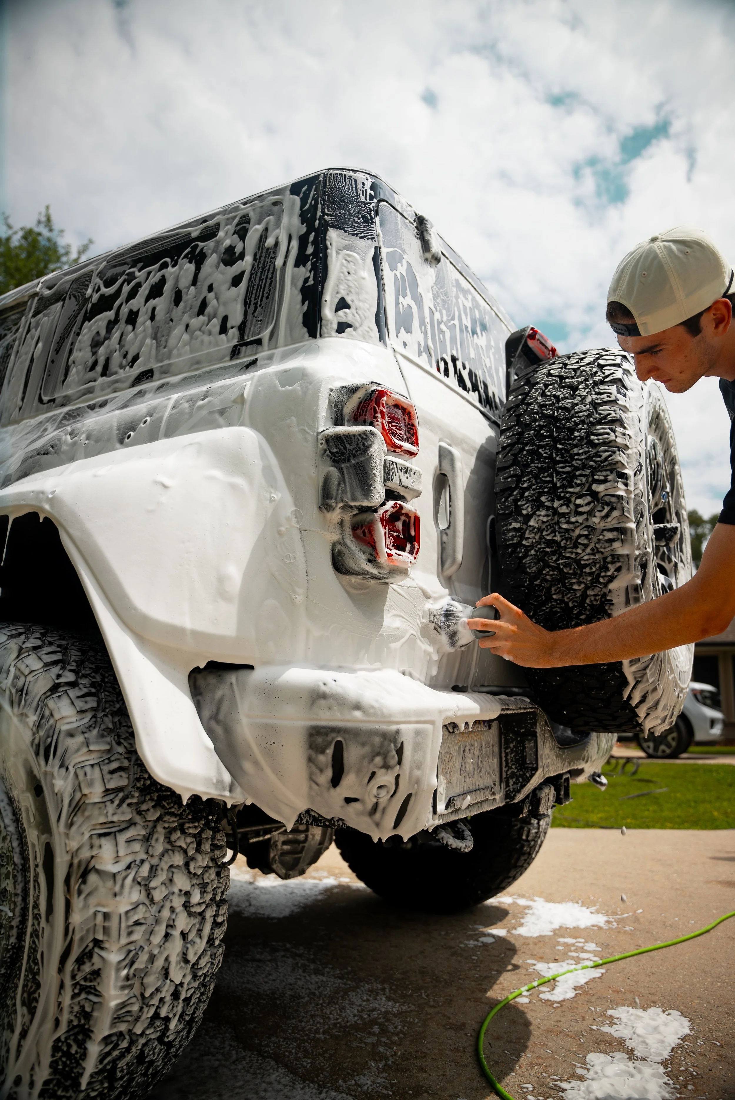 A person is washing a white off-road vehicle with soap suds and a foam nozzle. The vehicle's tires are covered in soap, and soap suds are visible on the ground.