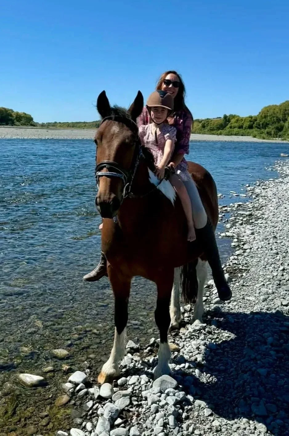 A woman and two young children riding a brown horse along a rocky shoreline next to a river under a clear blue sky.