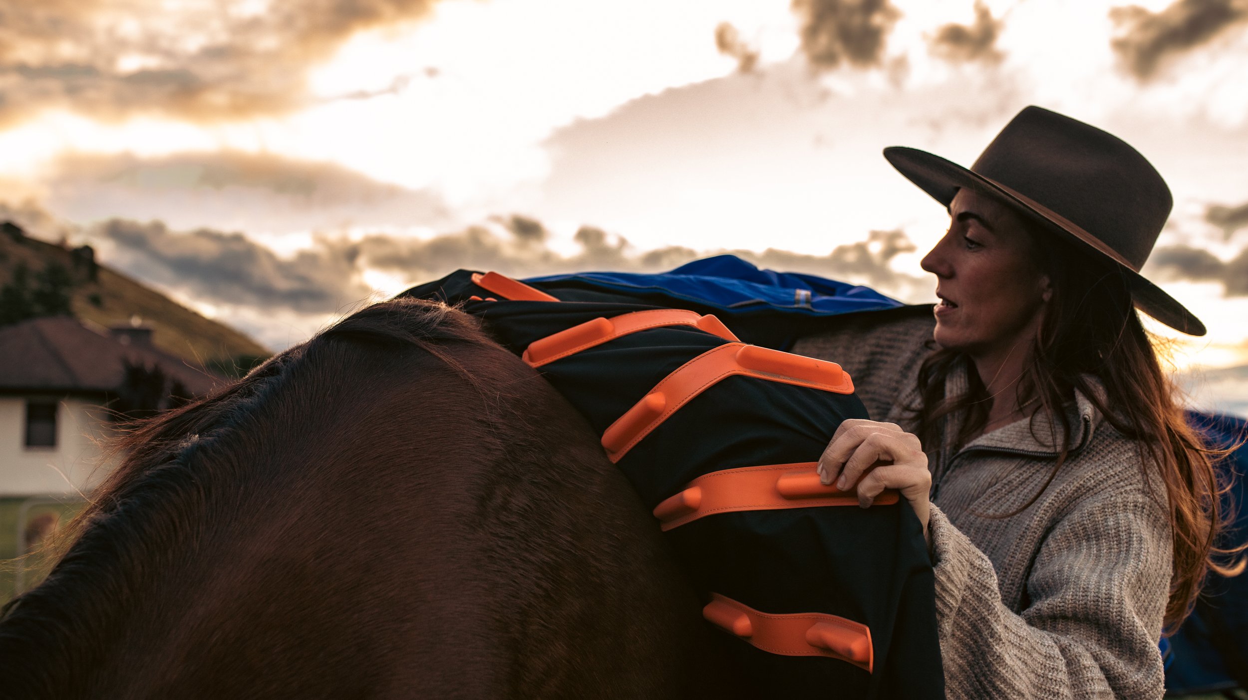 A woman wearing a wide-brimmed hat adjusting a horse cover on a horse during sunset.