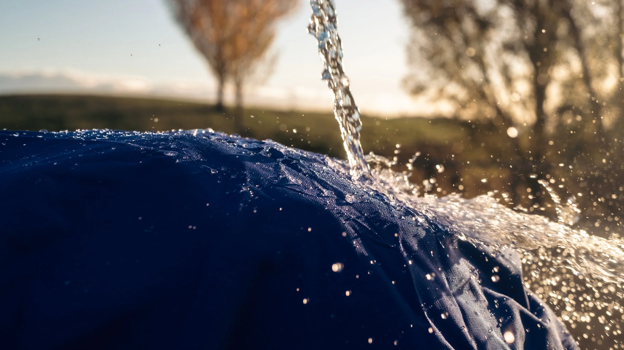 Close-up of water pouring over a blue waterproof horse cover fabric, with trees and sunlight in the background.