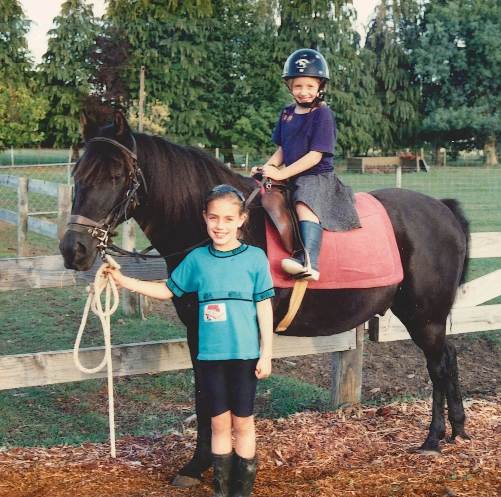 Two children at a horse riding arena with a brown horse. The girl riding the horse is smiling, wearing a black helmet, a blue T-shirt, and a gray skirt. The other girl standing in front is holding the horse's lead rope, smiling, wearing a blue T-shirt, black shorts, and rain boots, with a leash in hand. The background has trees and wooden fencing.