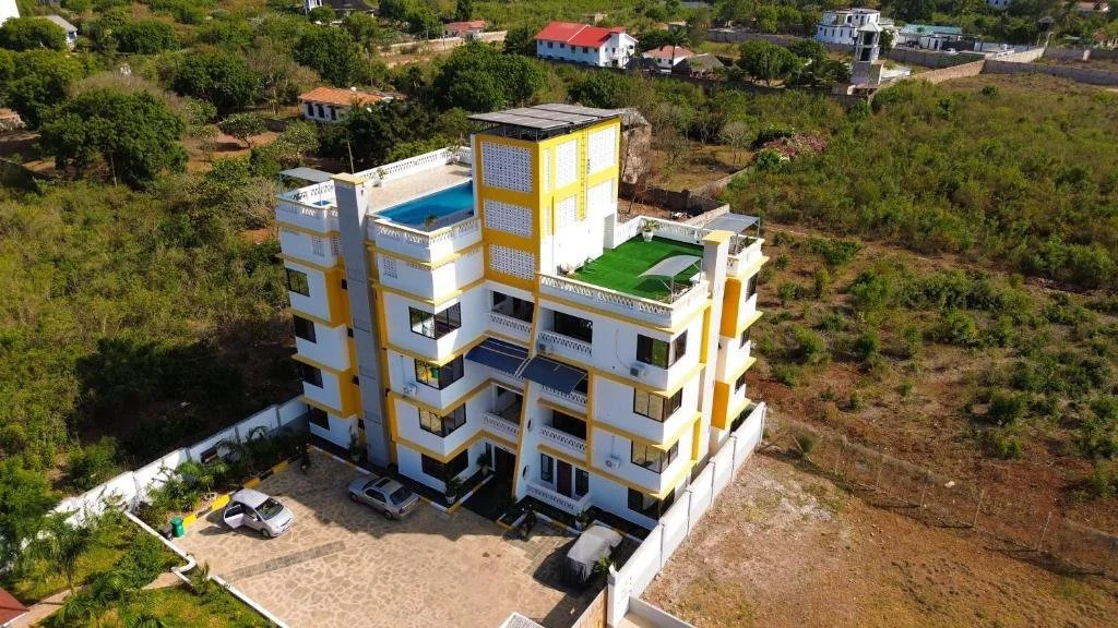 A multi-story white and yellow residential building with rooftop pools and green lawn, surrounded by a border wall, with parking area and neighboring houses in a green, semi-urban area.