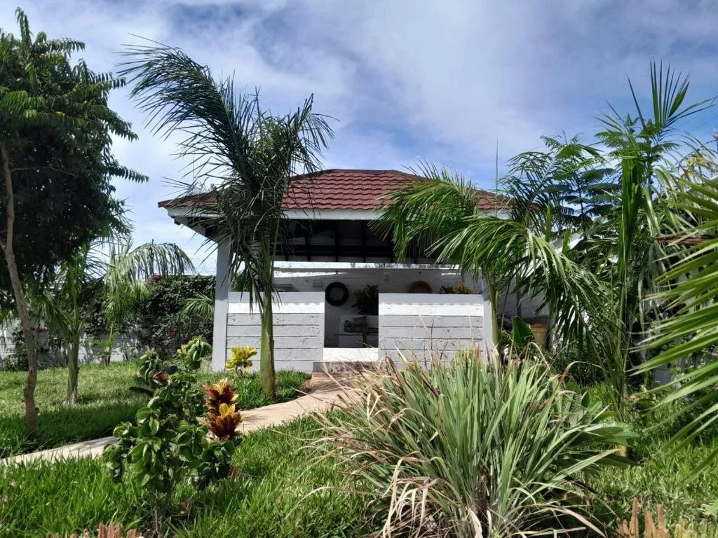 A small white house with a red tile roof surrounded by tropical plants and palm trees under a partly cloudy sky.