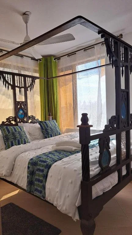 A bedroom with a large black wooden four-poster bed, white bedding, and patterned blue and green pillows. There are sheer curtains and bright green curtains, with sunlight coming through large windows. A ceiling fan is visible above.