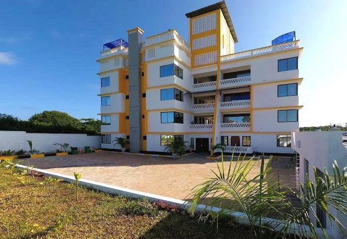 A modern multi-story residential building with white walls and yellow accents, surrounded by a paved courtyard and a garden area, under a clear blue sky.