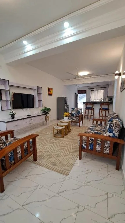 Living room with white walls, a large flat-screen TV on a white entertainment center, two wooden armchairs with patterned cushions, a sofa with matching cushions, a beige rug, and a small bar area with two stools in the background.