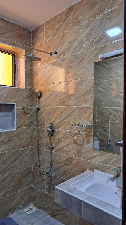 Bathroom shower area with large beige marble tiles, a small rectangular window, a wall-mounted showerhead, a handheld shower, a soap dish, a mirror, and a small white sink.