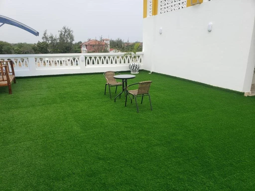 Rooftop terrace with artificial grass, a small table with two wicker chairs, a white decorative railing, and some potted plants, with houses and trees in the background.