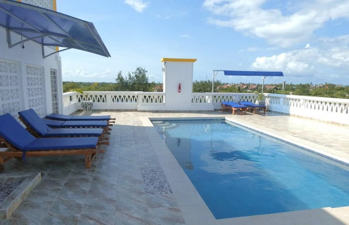 Rooftop swimming pool with blue lounge chairs, a large umbrella, and a scenic view of the sky and trees in the background.