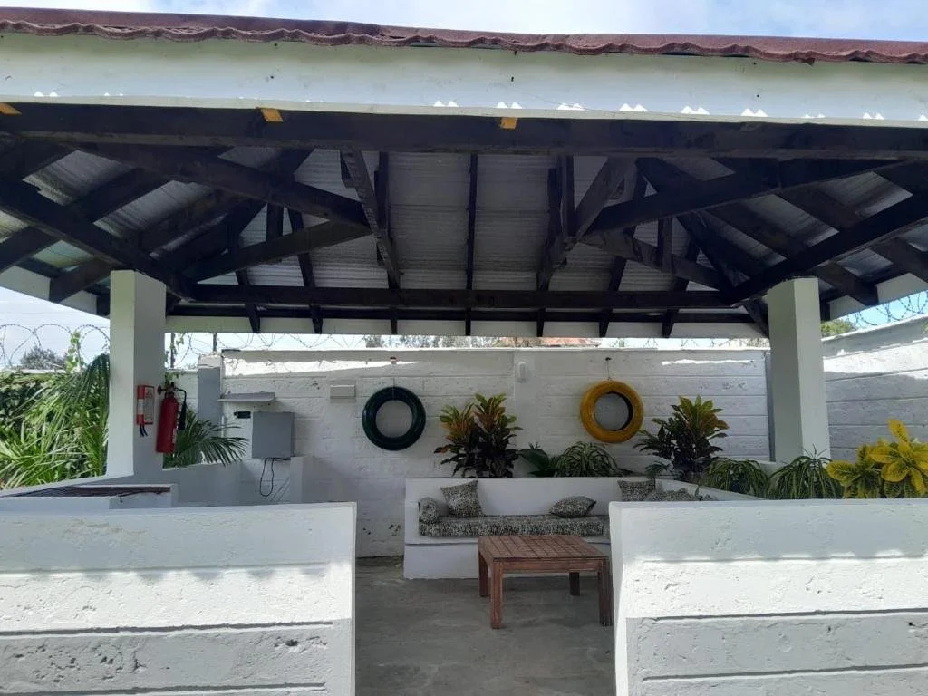 Outdoor covered patio with white walls, a wooden table, seating area with cushions, and decorative tires on the wall, surrounded by plants.
