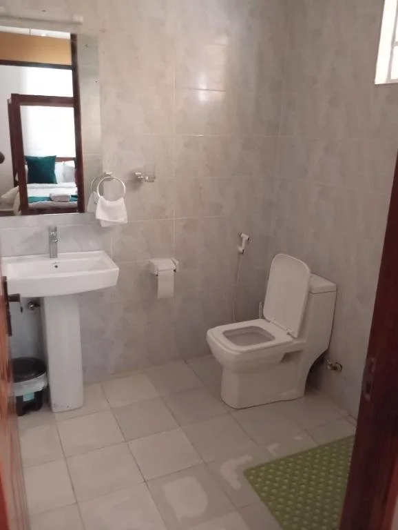 A small bathroom with light-colored tiled walls and floor, a white toilet, a white pedestal sink, a mirror above the sink, a toilet paper holder with a roll, a small green rug, and a window showing a glimpse of a bedroom with a bed and pillows.