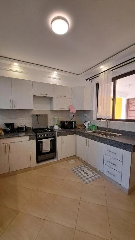 Kitchen with white cabinets, black countertop, stove, microwave, sink, and window with white curtains.